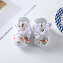 A pair of white baby girl shoes featuring a floral print, a white bow, and a heart-shaped accent.
