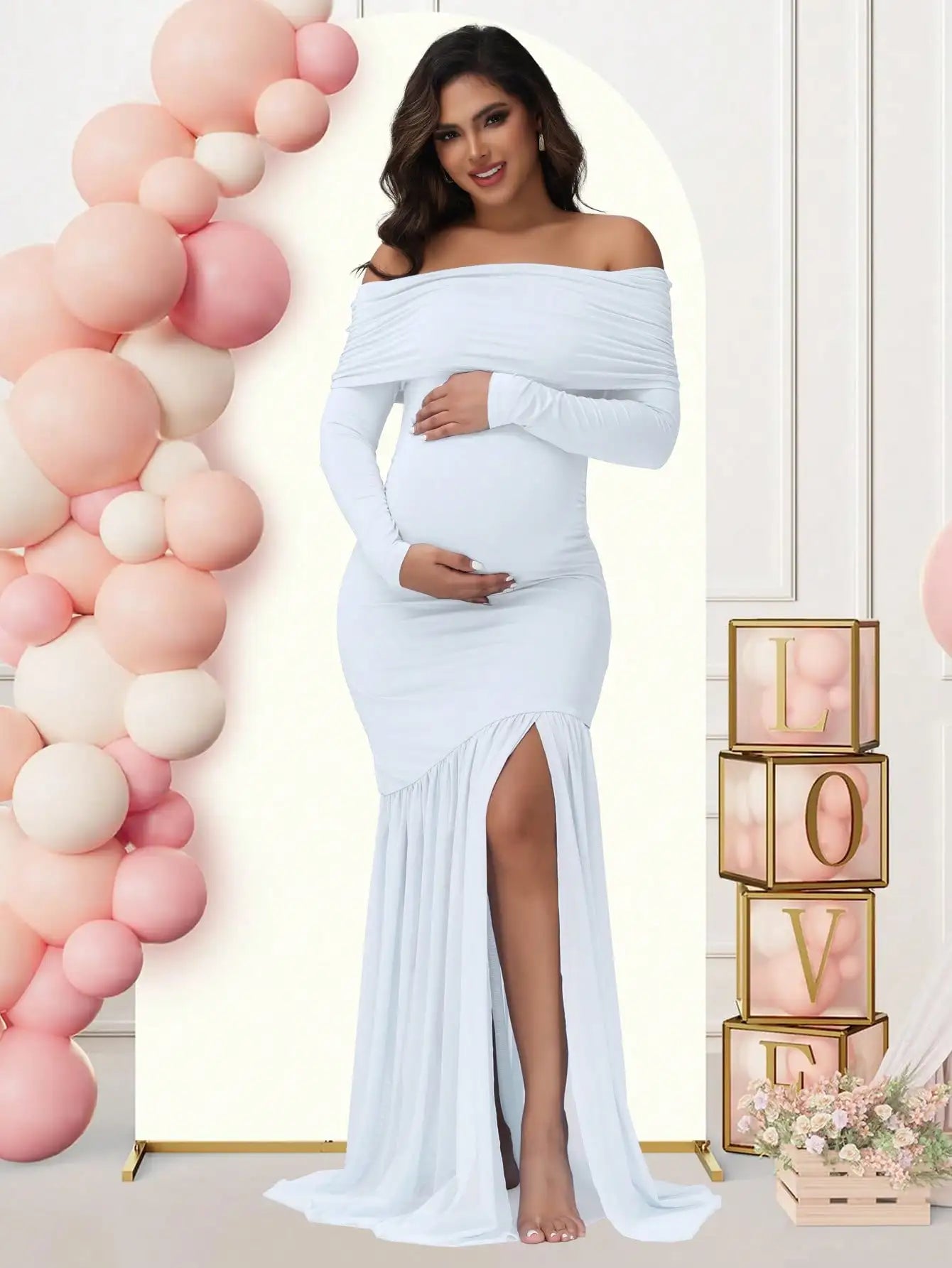 A pregnant woman wears a white off-shoulder long-sleeved maternity gown with a side slit.