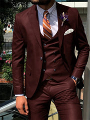 A man wears a dark burgundy three-piece suit with a white shirt and a striped orange tie.