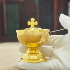 A gold-colored communion cup with a cross-topped lid and a matching spoon held in a gloved hand.
