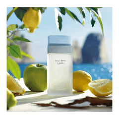 A bottle of Dolci and Bana Light Blue perfume sits on a surface surrounded by lemons and an apple.