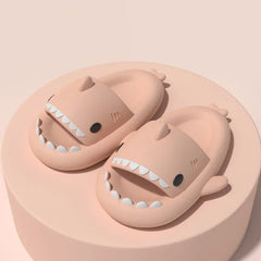 A pair of light pink shark-shaped slide sandals with white teeth on a matching circular platform.