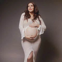 A pregnant woman wears a long-sleeve white sheer maternity dress with a front belly cutout.