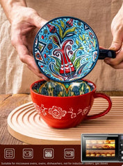 A person holds a colorful, patterned ceramic mug above a matching red mug on a wooden tray.