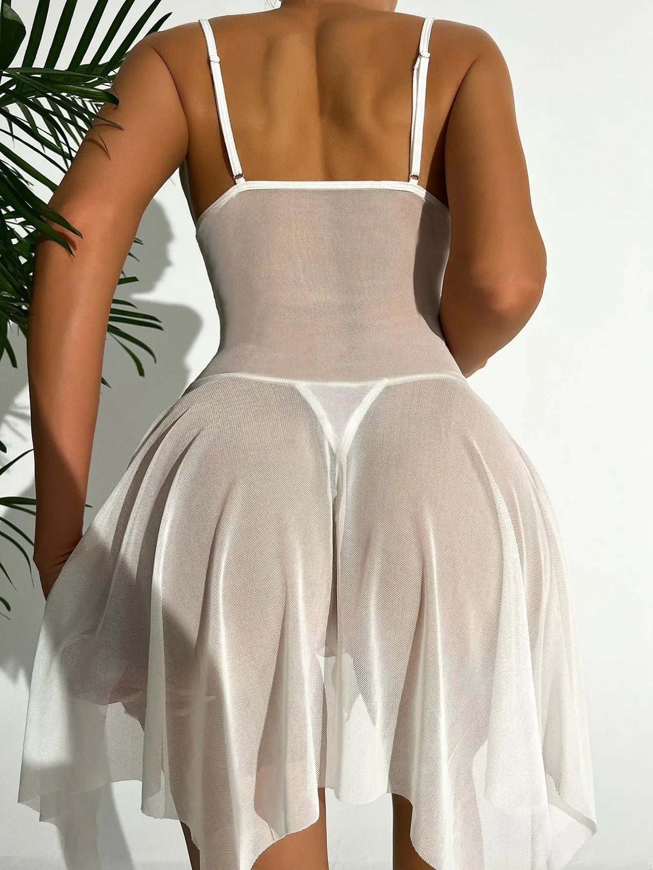 A woman is photographed from the back wearing the Sexy Lingerie Women Crotchless Sleepwear Summer Pajamas Lace Night Dress, a sheer white mini nightgown with visible undergarments; a green plant appears partially on the left.