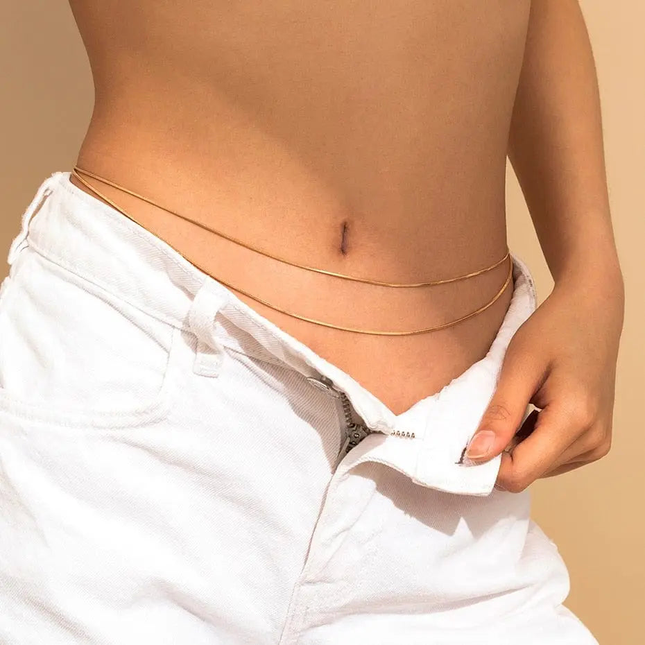 A person wears white jeans unbuttoned at the waist, revealing a Sexy Vintage Aesthetic Belly Chain Thin Beads Link Body Chain Waist Chain Belt Y2K Streetwear Summer Women Fashion Body Jewelry around their midriff against a beige background.