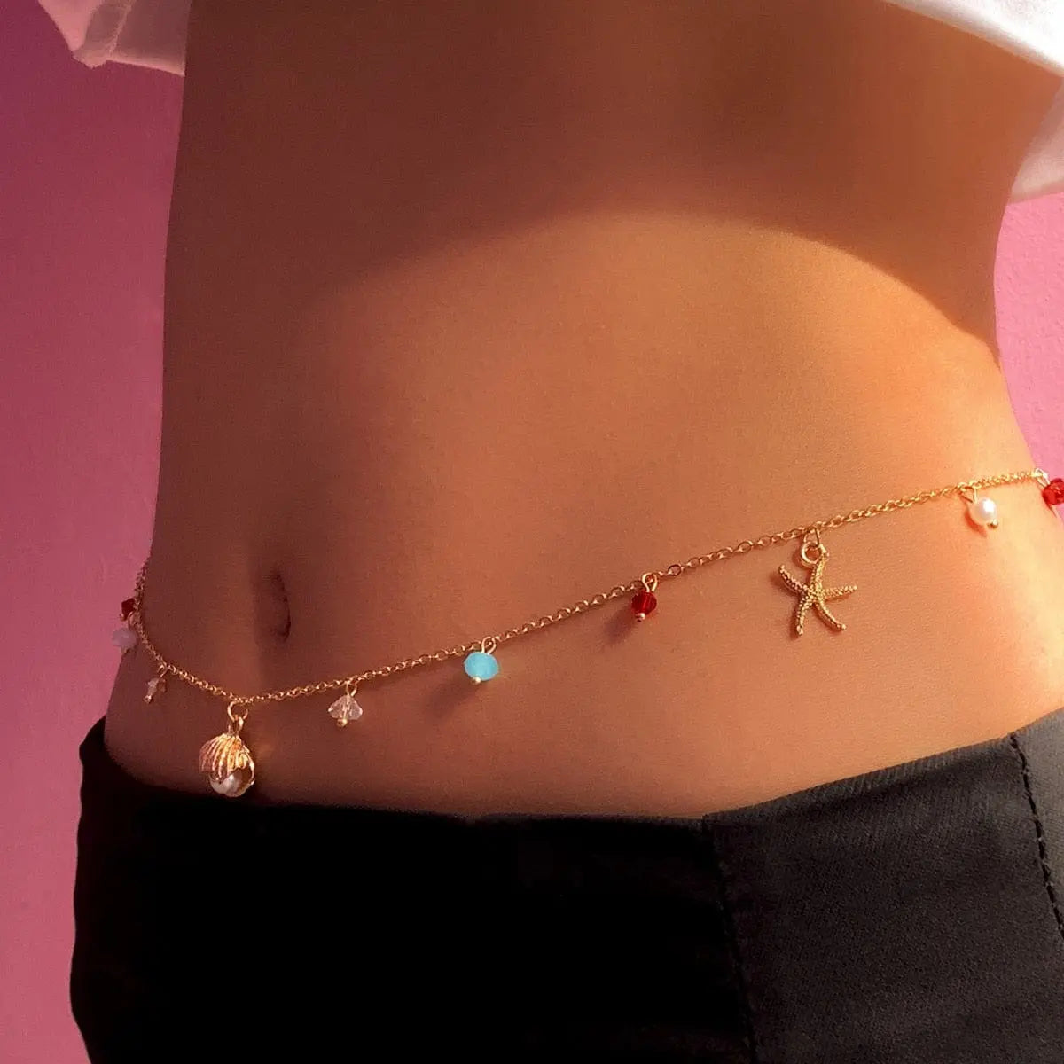 A close-up of a person wearing the Sexy Vintage Aesthetic Belly Chain Thin Beads Link Body Chain Waist Chain Belt—colorful beads and starfish charms—over black pants against a pink background.