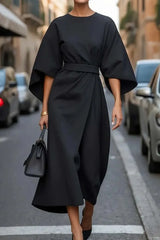 A woman wears an elegant black ankle-length dress with bat sleeves and a belted waist.