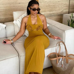 A woman wears a mustard yellow ankle-length halter dress featuring two large fabric floral accents.
