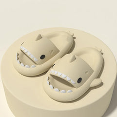 A pair of beige shark-shaped slide sandals with white teeth details resting on a circular platform.