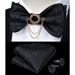 A black paisley bow tie set featuring a gold brooch with a chain, pocket square, and cufflinks.