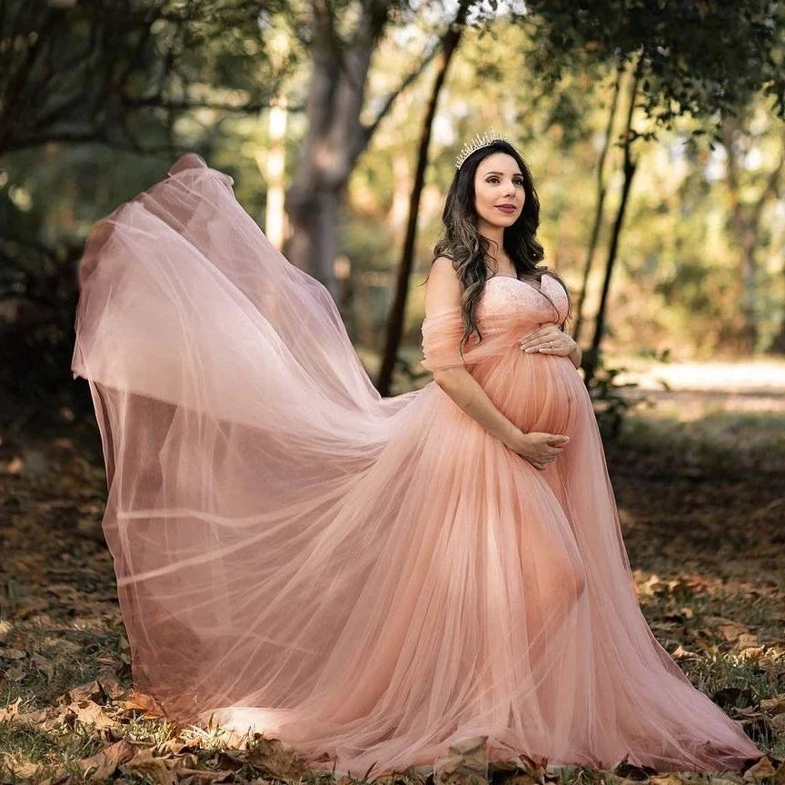 A pregnant woman wears a pink off-shoulder chiffon maternity gown with a flowing skirt.