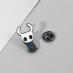 A metal enamel pin of the Hollow Knight character with a white mask and blue cloak.