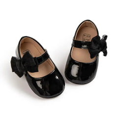 A pair of black patent leather baby shoes featuring a decorative bow and hook and loop closure.