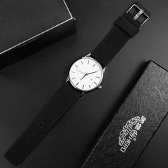 The Simple Men Quartz Watch, featuring a white face, thin black hour markers, and a black leather strap, is displayed diagonally on a dark surface beside two black watch boxes, one marked with Chinese characters.