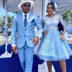 A man wears a sky blue two-button suit with a white hat and tie next to a woman in a blue dress.