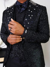 A man wears a black three-piece suit featuring a glittery finish and a beaded, diamond-studded lapel.