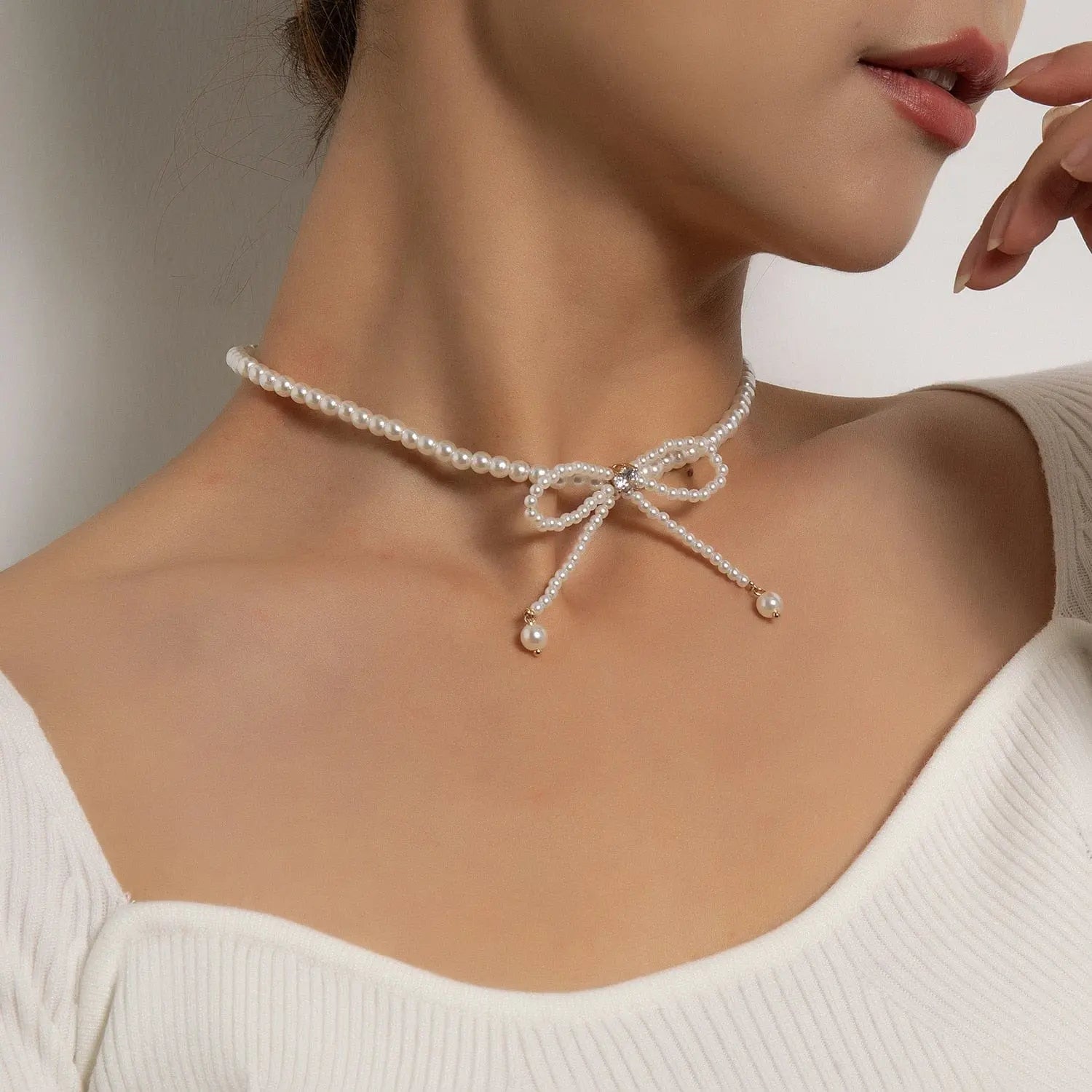 A woman wears the Vintage Style Simple 6MM Pearl Chain Choker Necklace, featuring a bow shape and two dangling pearl ends, paired with an off-shoulder white top.