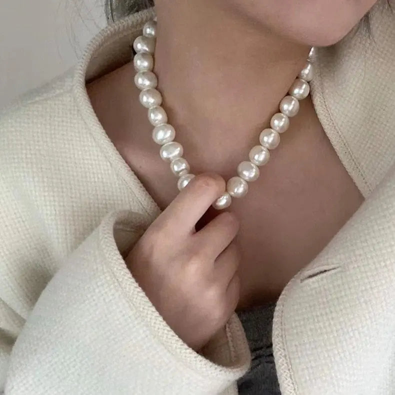 A woman wearing a cream textured jacket gently holds the Vintage Style Simple 6MM Pearl Chain Choker Necklace with shell pendant, highlighting the elegant fashion jewelry around her neck.