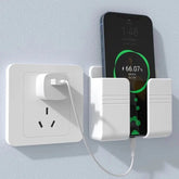 A white wall-mounted mobile phone holder holding a charging smartphone next to a wall outlet.