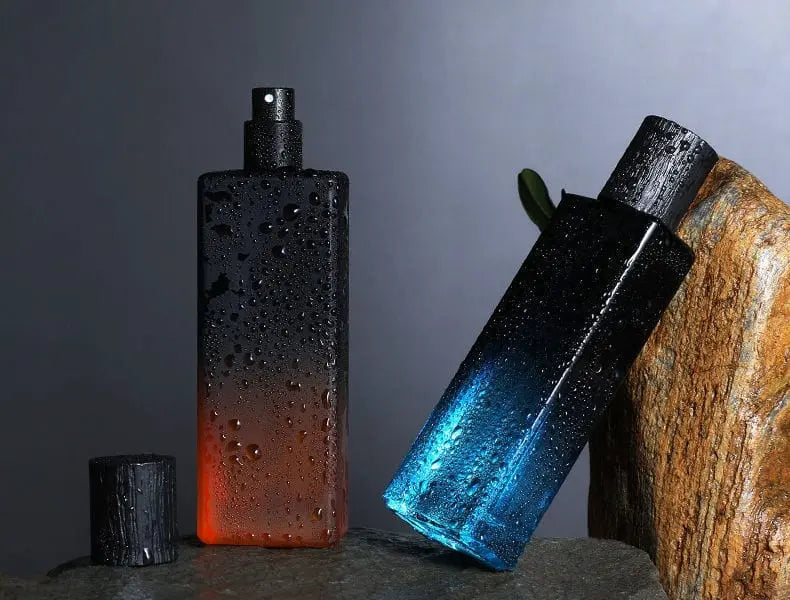 Two rectangular bottles of Wholesale perfume scents long lasting fragrances men cologne fragrance—one upright in an orange-black gradient with water droplets, the other tilted in blue-black—are displayed on stone surfaces against a gray background.