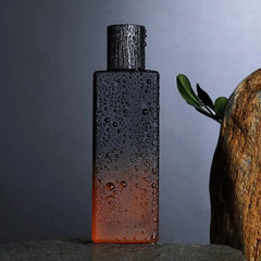 A rectangular bottle of Wholesale perfume scents long lasting fragrances men cologne stands upright, fading from black to orange, topped with a dark cap. Water droplets cover it, with a leafy branch and textured rock in the background.