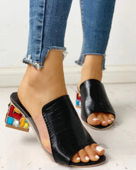 Black croc-embossed slide sandals featuring a clear PVC side panel and a chunky heel with colorful rhinestones.