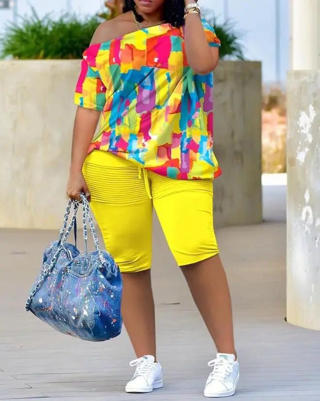 A woman models the Womens Two Piece Sets Outfit Melting Colors All Over Print Skew Neck Top & Shorts Set, styled with white sneakers and a large blue splatter tote. Her face is not shown.