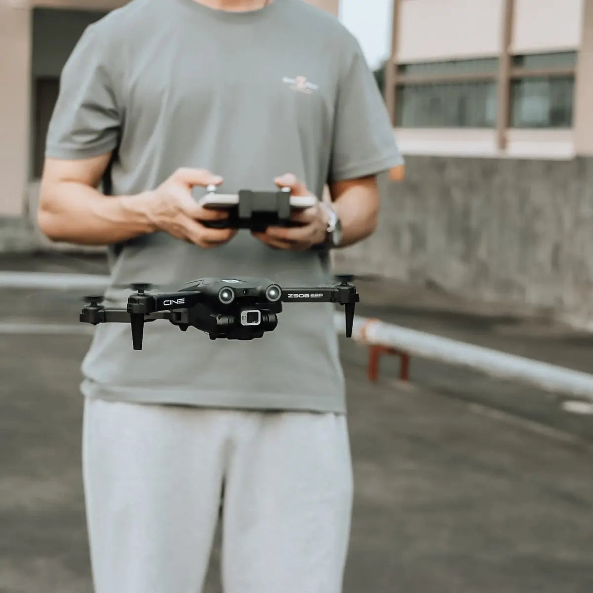 A person in a gray t-shirt and light pants operates the Z908 Pro Drone Professional 4K HD Camera Quadcopter with a controller outdoors on pavement near a building. This toy features three-sided obstacle avoidance for safer flights.