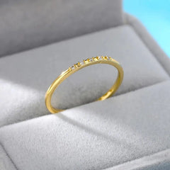 The Zircon Engagement Wedding Ring for women features small, evenly spaced cubic zirconia stones set in gold-color stainless steel, displayed on a light gray velvet jewelry box with a soft blue background.