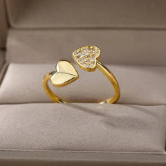 A gold color stainless steel ring for women, featuring heart-shaped ends—one solid, one with sparkling cubic zirconia—displayed on a beige jewelry box. Perfect as an engagement or wedding ring. 2023 design, free shipping.