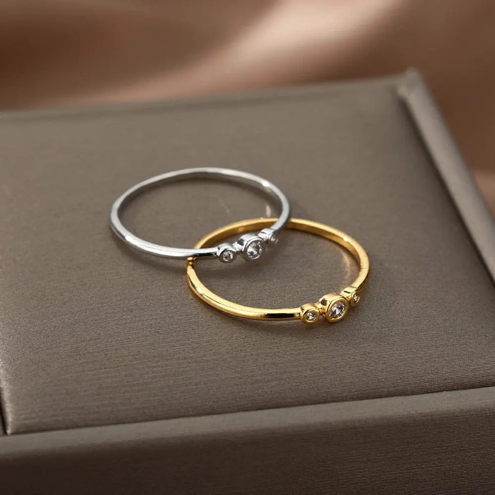 Two elegant gold color stainless steel zircon engagement rings for women, each featuring three sparkling cubic zirconia stones, are displayed on a soft brown jewelry box with a satin fabric background.