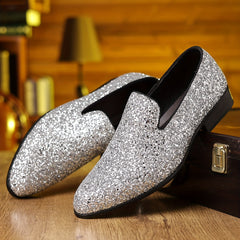 A pair of silver glittery slip-on formal shoes with a pointed toe resting on a wooden surface.
