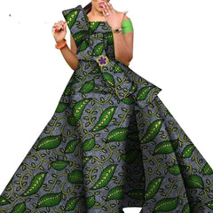 A woman wears a floor-length African print dress featuring a green leaf pattern on a grey background with a decorative bow detail.
