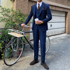 A man wears a navy blue wool-blend three-piece suit with a herringbone pattern and brown loafers.