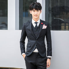 A man wears a slim-fit black three-piece suit with a white shirt, black tie, and a red lapel pin.
