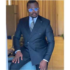 A man wears a black double-breasted suit with a patterned tie and blue-tinted sunglasses.