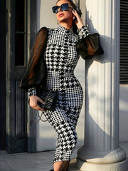 A woman wears a fitted, high-neck houndstooth dress featuring long, sheer black mesh sleeves.