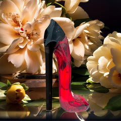A red stiletto-shaped perfume bottle stands in front of large, pale flowers.
