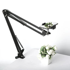 A black adjustable overhead desk mount holding a smartphone above a small potted flower arrangement.