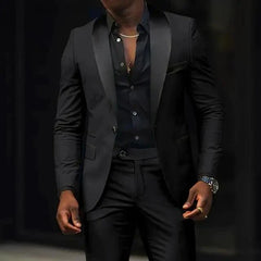 A man wears a black slim-fit two-piece suit featuring a shawl lapel blazer and matching trousers.