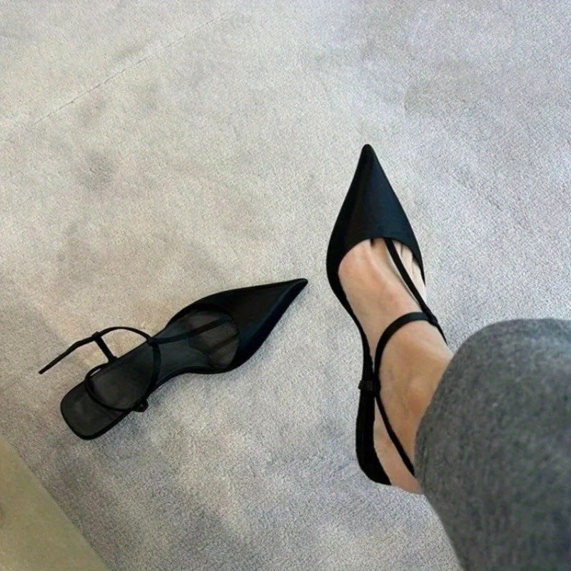 A pair of black pointed-toe stiletto sandals with thin ankle straps worn on a light surface.