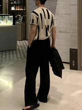 A man wears a short-sleeve, cream-colored knitted sweater featuring a black abstract block pattern.