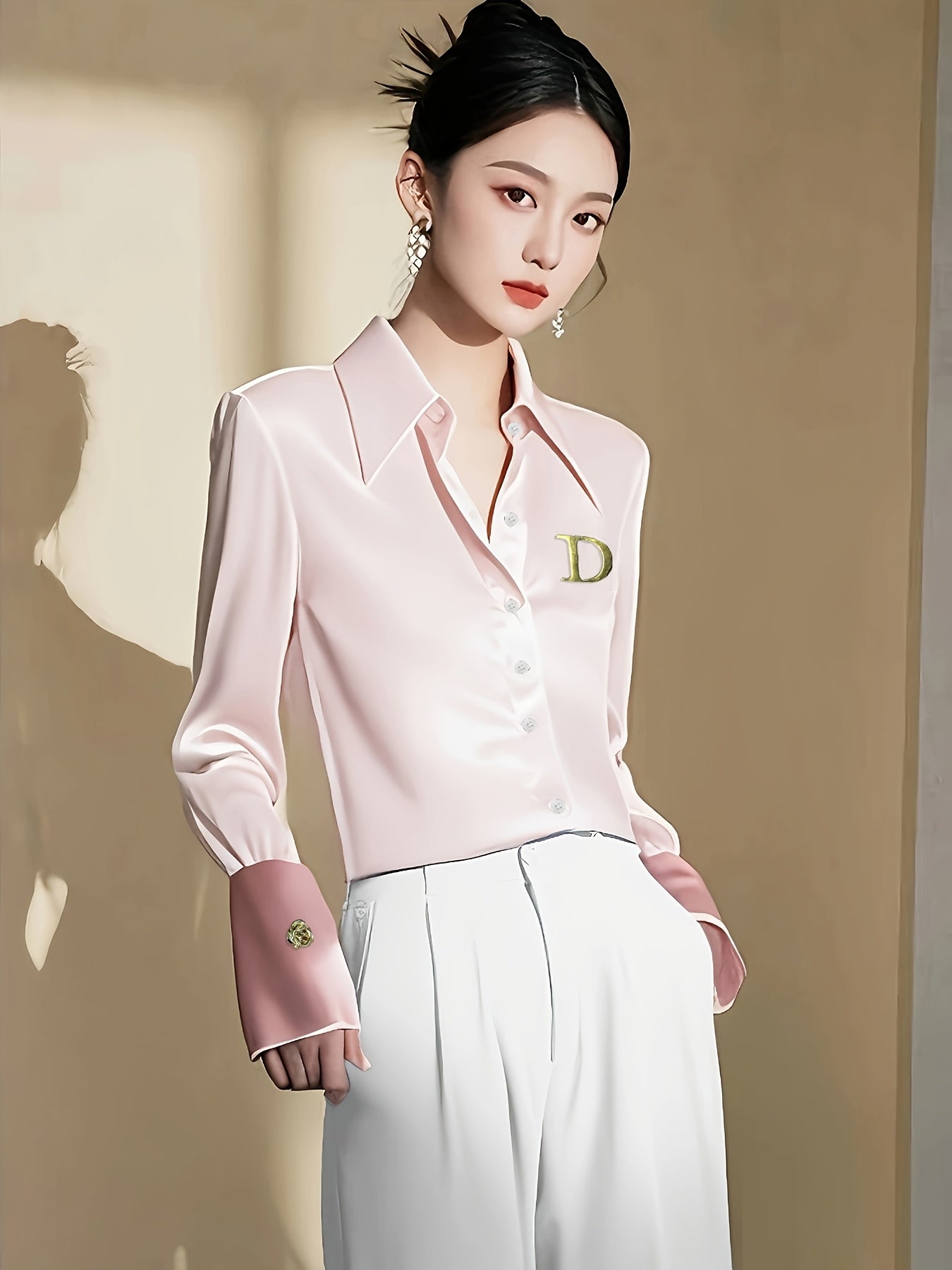 A woman wears a pale pink long-sleeved formal shirt with a wide collar and a gold D logo.