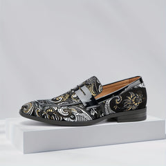 A black slip-on loafer with intricate white and gold floral embroidery on a white display block.