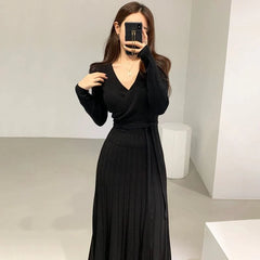 A woman wears a long-sleeved black V-neck knit dress with a pleated skirt and a waist-cinching tie.
