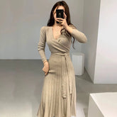 A woman wears a beige long-sleeved V-neck knit dress with a pleated skirt and a waist tie.