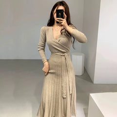 A woman wears a beige long-sleeved V-neck knit dress with a pleated skirt and a waist tie.