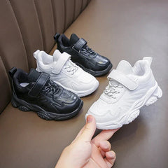 Four children's sneakers in black and white with hook and loop closures on a brown surface.