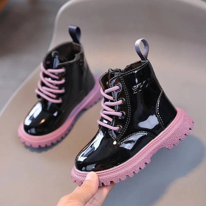 A pair of shiny black ankle boots for children with pink laces and thick pink rubber soles.